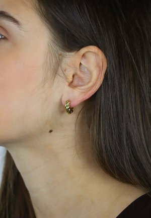 Złota kolczyk kołowy z oplecionym wzorem, prezentujący polerowaną teksturę. Noszony na płatku ucha, na tle długich, ciemnych włosów.