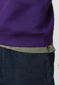 Sweat-shirt violet avec ourlet et col côtelés, superposé à une chemise verte. Jean en denim foncé avec des détails de couture jaunes.