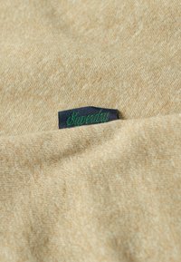 Tessuto beige con una piccola etichetta blu scuro con scritta verde ricamata "Superdry" applicata alla cucitura.