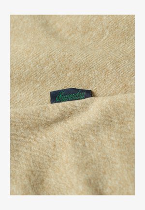 Tela texturizada beige con un patrón sutil, con una etiqueta azul oscura que tiene el texto "Superdry" en verde claramente visible.