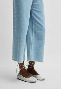 Ljusgrå platta skor med rund tå, mjukt lädermaterial och snören som viras runt ankeln, kombinerat med avkortade blå denimjeans.