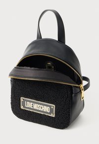 Mini nahrbtnik Black Love Moschino z teksturiranim sprednjim žepom, zlatim zadrgo in ročajem na vrhu, delno raztegnjen, ki razkriva podlogo znotraj.