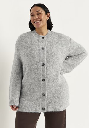 Kvinde smilende, iført en blød, lysegrå cardigan med knapper og mørke bukser, stående med den ene hånd på hoften mod en ensfarvet baggrund.