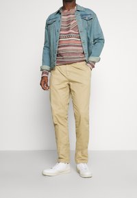 Giacca di denim blu sopra un maglione a maglia con motivo, chinos color khaki e sneaker bianche. I dettagli notevoli includono il design geometrico del maglione.