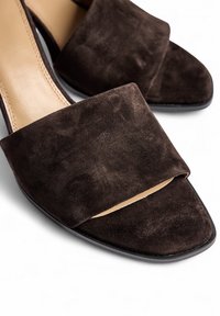 Sandales à talon plat en suède marron avec une large bride supérieure et un bout ouvert. Semelle plate et design simple, mettant en avant une texture douce et des lignes épurées.