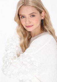 Weißer Strickpullover mit strukturierten Ärmeln, die mit runden Pailletten verziert sind. Das Model hat hellblonde Haare, minimales Makeup und schwarze Ohrringe.
