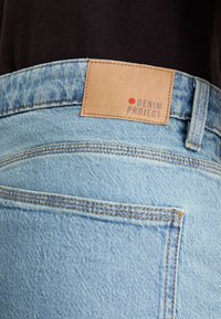 Svijetloplave traper hlače s istaknutom stražnjom džepom. Upotpunjene svijetlosmeđom etiketom s utisnutim natpisom "DENIM PROJECT" i crvenom točkom.