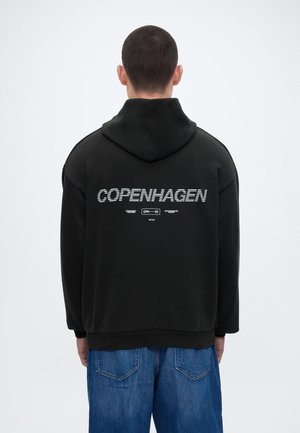 Άνδρας που φοράει μαύρο φούτερ με κουκούλα με το κείμενο "COPENHAGEN" στην πλάτη, συνδυασμένο με μπλε τζιν, κοιτάζει ένα λευκό φόντο.