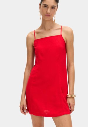 Frau trägt ein rotes ärmelloses Minikleid mit dünnen Trägern, goldene Creolen und ein massives goldfarbenes Armband am rechten Handgelenk.