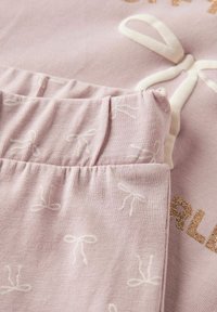 Gaiši rozā kokvilnas apģērbs ar teksturētu baltu lentīšu loku rakstu. Satur zelta gliteri iespiestu detaļu. Mīksta, elastīga josta.