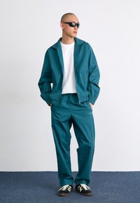 Survêtement teal avec une veste zippée et un pantalon à taille élastique. Comprend un t-shirt blanc en dessous et des baskets noires avec des lacets blancs.