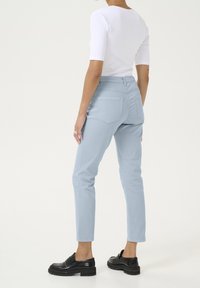 Pantalon en denim bleu clair à coupe slim, avec des poches arrière et des coutures contrastées, associé à un haut blanc à manches longues et des chaussures noires.