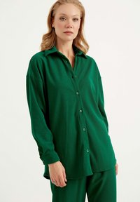 Grüne Button-Down-Bluse mit lockerer Passform, aus strukturiertem Stoff. Verfügt über einen Kragen, lange Ärmel und einen abgerundeten Saum mit Knopfdetails.