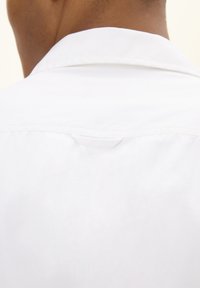 Witte overhemdd, gemaakt van een gladde stof, met een kleine stoffen lus aan de achterkant van de hals om op te hangen. Naaddetails zijn zichtbaar.