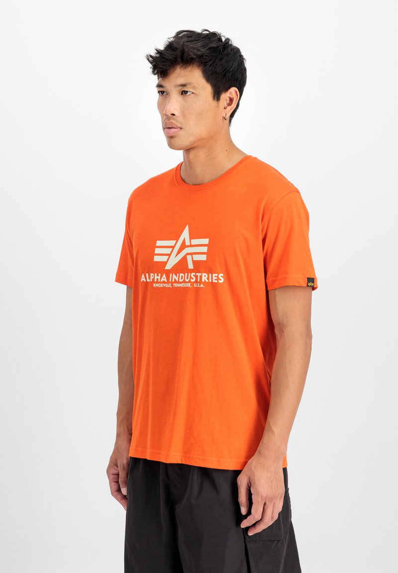 Oranje katoenen T-shirt met een ronde hals, met een witte logo-ontwerp "ALPHA INDUSTRIES" prominent weergegeven op de voorkant.