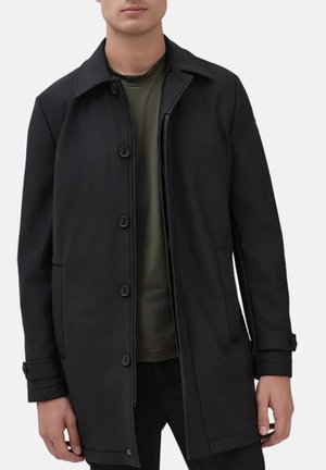 Cappotto nero con colletto strutturato, zip frontale e chiusura a pulsante. Presenta tasche laterali e una texture liscia. Indossato sopra un top verde scuro.