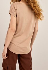 Beige short-sleeve t-shirt with rolled cuffs, made of soft fabric, featuring a curved hemline and relaxed fit. Brown cargo pants visible.