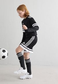 Maillot de sport noir avec des rayures blanches, shorts noirs assortis, chaussures de soccer blanches, et chaussettes noires hautes avec des rayures grises. Un ballon de soccer à proximité.