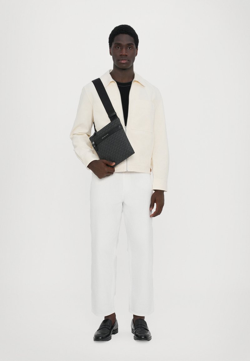 Sac bandoulière noir avec motif texturé, porté par une personne en veste crème, chemise noire, pantalon blanc et mocassins noirs. Fond uni.