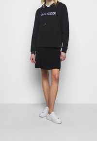 Černá mikina s logem "Ochil Hugo", v kombinaci s černou sukní. Modelka má na sobě bílé tenisky. Hladký materiál, ležérní design a volný střih.