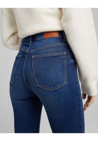 Dámské modré denimové džíny s vysokým pasem, se dvěma zadními kapsami, kontrastním šitím a hnědou koženou etiketou na pasu. Měkká struktura.