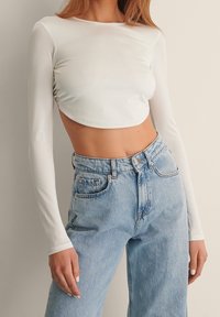Weißes langärmliges Crop Top mit glatter Textur und gerafftem Seiten-Detail, kombiniert mit hochgeschnittenen hellblauen Denim-Jeans.