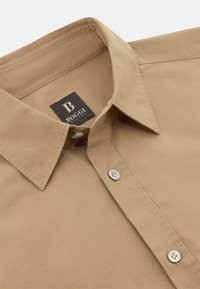 Hnědé košile s rozepínacím límcem, s jemnou texturou a diskrétním prošíváním. Obsahuje štítek s logem a knoflíkové detaily.