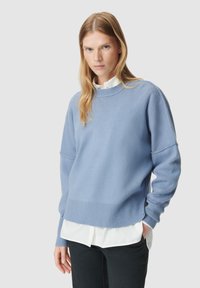 lichtblauwe gebreide trui met een ribbenhalslijn, verlaagde schouders en een relaxte pasvorm, gelaagd over een wit overhemd met kraag.