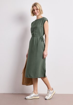 Frau trägt ein ärmelloses olivgrünes Midikleid und silberne Sneaker, hält eine große braune Tragetasche und geht an einer weißen Wand entlang.