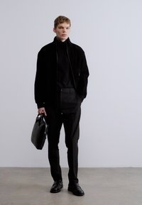 Čierna semišová bunda, čierny rolák a nohavice. Jemná textúra. Čierna kožená taška. Model stojí pred svetlým pozadím.