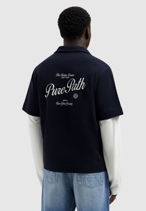 Person, die ein marineblaues Kurzarmhemd über einem weißen Langarmshirt trägt, mit der Aufschrift "Pure Path" und Text auf der Rückseite des Hemdes.