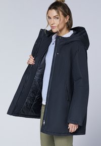Schwarze Kapuzenjacke mit Reißverschluss und Druckknopfverschluss, mit Seitentaschen und gefüttertem Innenraum. Wird über einem hellblauen Pullover getragen.
