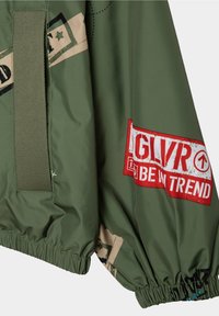 Giacca verde in nylon con polsini elasticizzati, grandi patch grafiche rossi e beige, e un dettaglio a strappo testurizzato.
