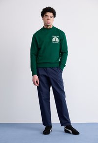 Groene sweatshirt met logo op de voorkant; gecombineerd met marineblauwe broek en zwarte sneakers. Soepele stof met ribbels aan de manchetten en de zoom.