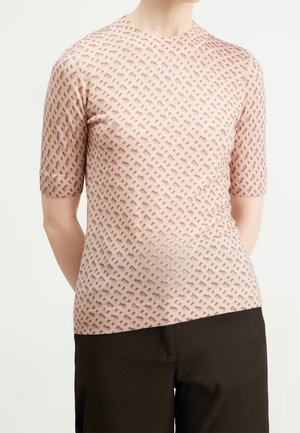 Personne portant un haut moulant à manches courtes de couleur rose clair avec un motif géométrique texturé et un pantalon noir, debout les mains derrière le dos.