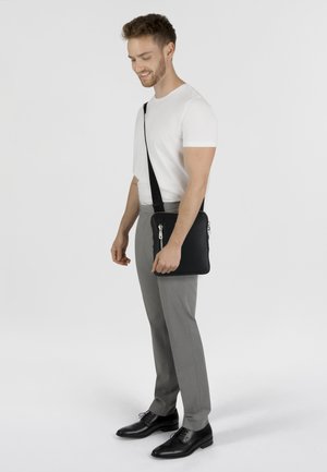 Borsa a messanger nera in pelle con superficie testurizzata, cinturino regolabile e cerniere argentate, tenuta da una persona che indossa pantaloni grigi e una camicia bianca.