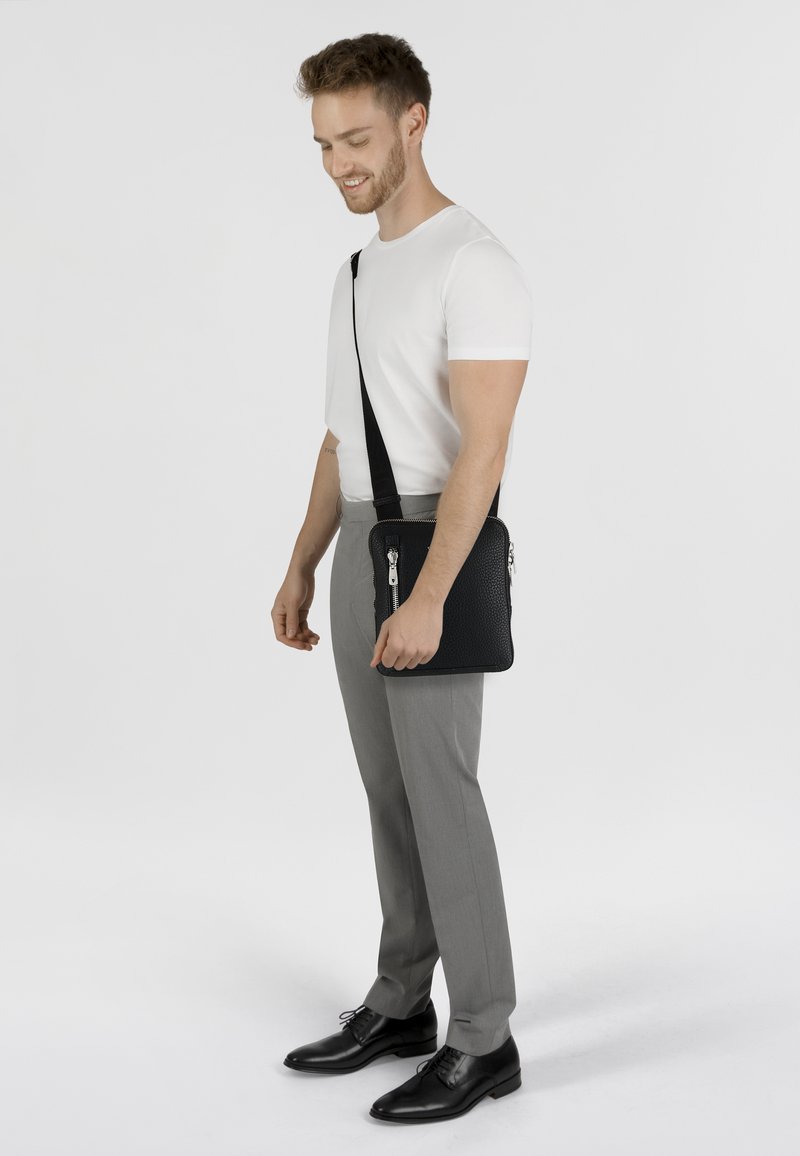 Bandolera de cuero negro con superficie texturizada, correa ajustable y cremalleras plateadas, sostenida por una persona que lleva pantalones grises y una camisa blanca.