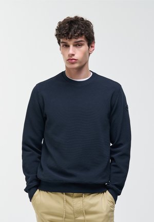 Jonge man met donker krullend haar, gekleed in een marineblauwe trui en beige broek, staand tegen een effen lichte achtergrond.