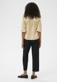 Satin-beige top med korte puffærmer, parret med sorte bukser og sorte loafers, set fra ryggen. Glat tekstur og skræddersyet pasform.