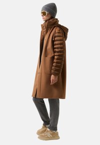 Hnědý kabát s nadýchanými rukávy, šedá pletená čepice a oversized sluneční brýle, spárované se šedými kalhotami a béžovými teniskami.