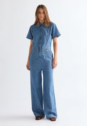 Žena oblečená v modrom denimovom overal s krátkym rukávom, širokými nohavicami a prednými vreckami, stojaca pred jednoduchým svetlým pozadím.