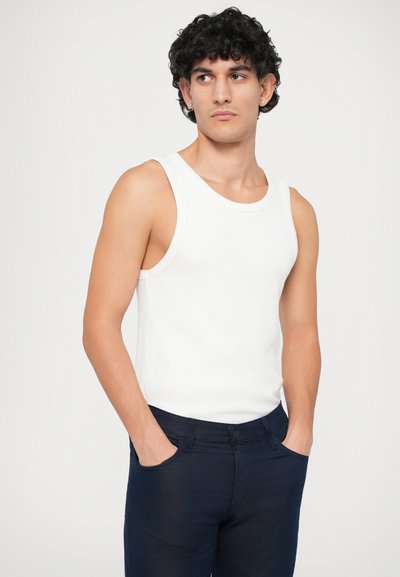 Camisola branca canelada com grandes aberturas para os braços, combinada com calças justas azul marinho. Textura suave e design simples e clássico.