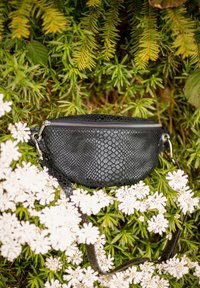 Sort taske med slangeskindsprint og sølv lynlås, oval form, justerbar rem, der hviler på grønne planter og hvide blomster. Tekstureret lædermateriale.