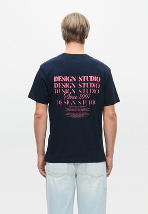T-shirt en coton bleu marine avec un texte rose vif au dos : "STUDIO DE DESIGN," "Depuis 2007," et un texte descriptif supplémentaire. Manches courtes, coupe décontractée.