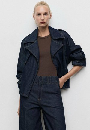 Tumman denim-takki, jossa on leveät kaulukset ja käännetyt hihat, yhdistettynä ruskeaan tiukkaan paitaan ja korkeavyötäröisiin, löysiin tumman denim-housuihin.