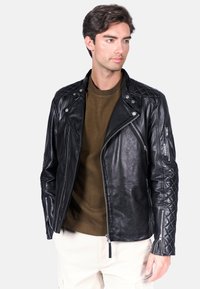 Czarna skórzana kurtka motocyklowa z pikowanymi detalami na rękawach, srebrnymi zamkami i dopasowanym krojem, noszona na oliwkowej koszuli.