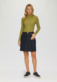 Groene lange mouw top met verticale naden, gecombineerd met een donkerdenim geplooide rok met voorzakken en contrasterende stiksels. Zwarte sneakers.