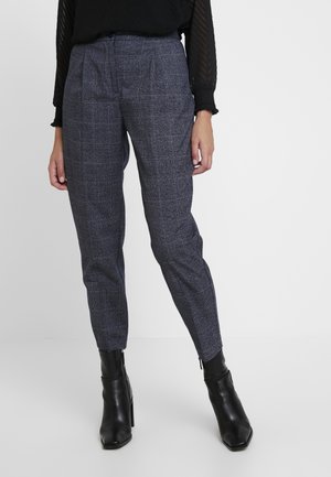 Femme portant un pantalon gris à carreaux ajusté et des bottines noires, debout devant un fond blanc uni.