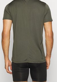 Oliivinvihreä, lyhythihainen urheilupaita, jossa on kapea istuvuus, sileä pinta ja yksinkertainen muotoilu, yhdistettynä mustiin urheilushortseihin.