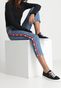 Jeans blu a vita alta con strisce laterali rosse e nere, abbinati a sneakers nere con lacci bianchi e una tomaia in tela testurizzata.